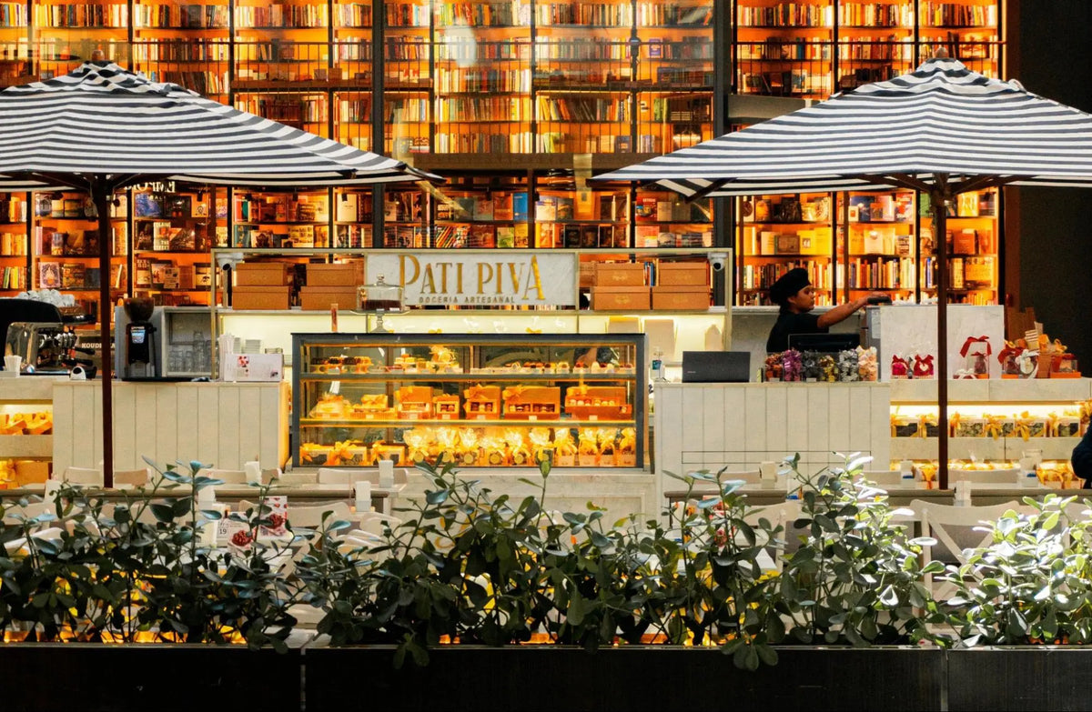 Fachada de um quiosque da Pati Piva com vitrines recheadas de doces e grandes ombrelones listrados em preto e branco. Ao fundo, destaca-se uma imensa estante de livraria iluminada repleta de livros do chão ao teto.