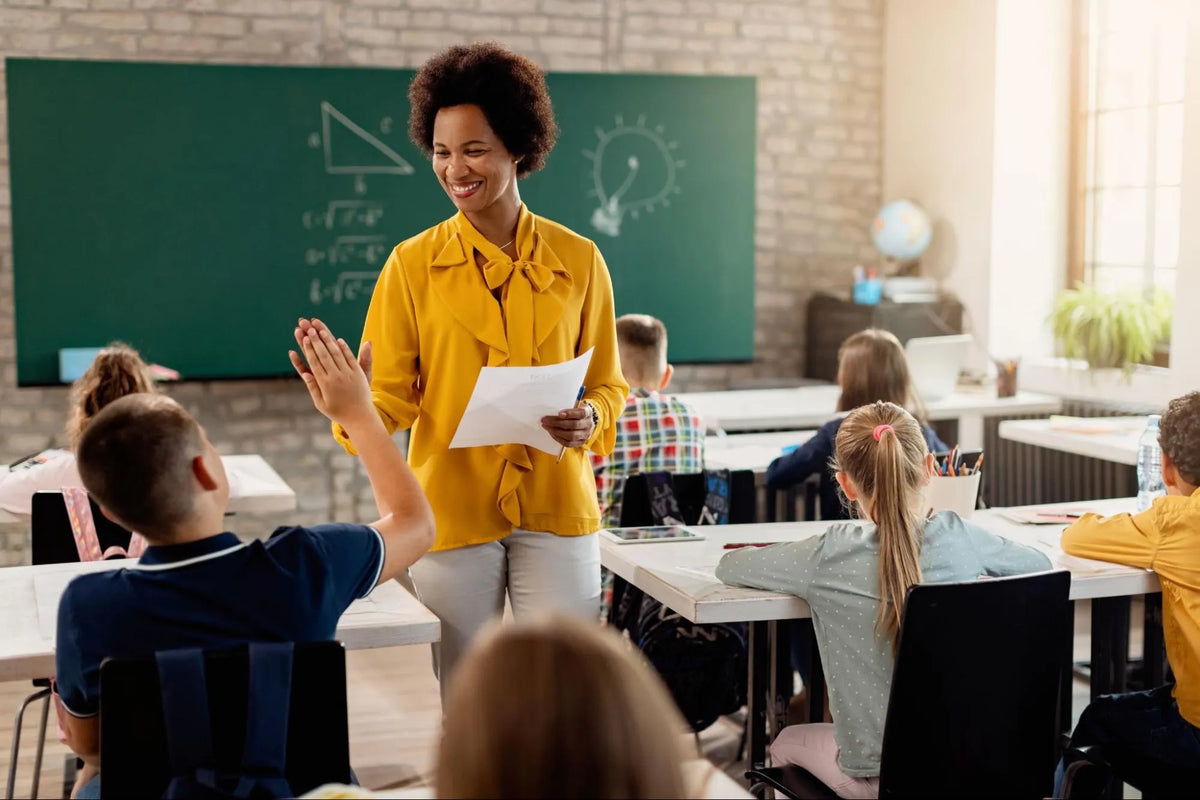 Professora sorridente vestindo blusa amarela cumprimentando um aluno com um "high five" em sala de aula iluminada. Em segundo plano, outros estudantes trabalham em suas carteiras diante de um quadro verde com anotações.